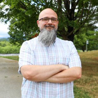 Professional headshot of Jeremy Bise, a man with a shaved head, glasses, and a long gray beard, wearing a blue and white plaid button-down shirt, standing outdoors with trees in the background.