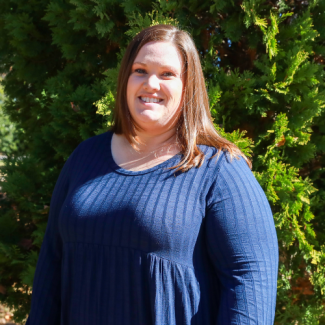 Jessica Jennings, smiling in front of green foliage, wearing a navy blue ribbed top.