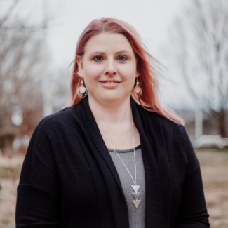 Jessie O'Quinn, smiling with pink hair, wearing a black cardigan over a grey top and a silver geometric necklace.