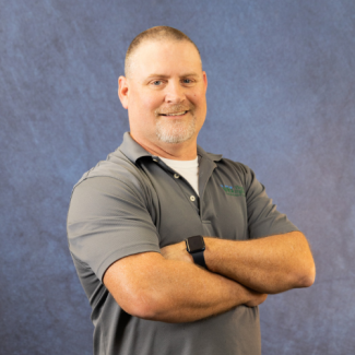 Johnnie Keene, smiling with arms crossed, wearing a grey polo shirt with a VHCC logo.