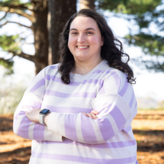A woman with long wavy dark hair smiles with her arms crossed. She is wearing a cream-colored sweater with light purple horizontal stripes and a silver smartwatch. Tall trees and a bright, soft-focus landscape are in the background.
