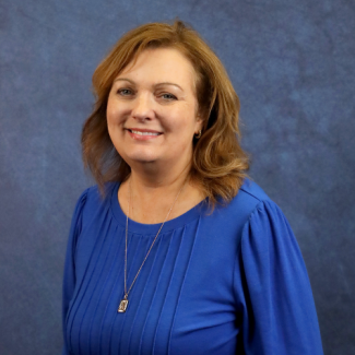 A woman with shoulder-length reddish-brown hair smiles against a blue textured backdrop. She is wearing a bright blue pleated blouse and a gold pendant necklace.