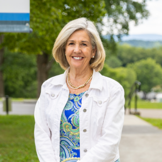 A smiling woman with straight, blonde, chin-length hair, wearing a white denim jacket over a blue, green, and yellow paisley print dress, accessorized with gold necklaces. She is standing outdoors with a soft-focus background of trees.