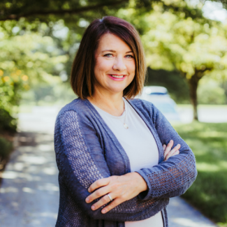 Professional headshot of Karen Copenhaver, a smiling woman with shoulder-length brown hair, wearing a white top and a blue open-knit cardigan, standing in a sunlit outdoor setting with trees.