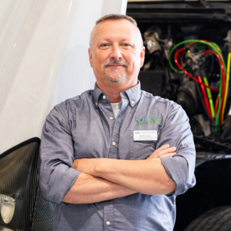 A man with a short grey beard and glasses smiles against a blue textured backdrop. He is wearing a grey quarter-zip pullover sweater over a light-colored collared shirt.