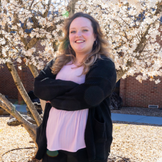 Portrait of Kelli Peck, a woman with long brown hair, smiling with arms crossed in front of a blooming white tree.
