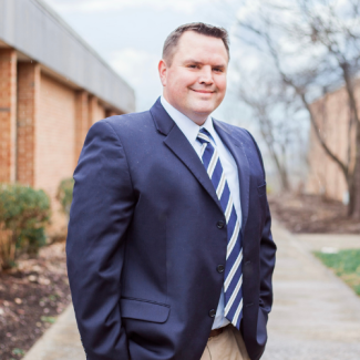 Headshot of Kevin Widener, a man with short brown hair, smiling and wearing a navy blue suit jacket with a striped tie. He is standing on an outdoor walkway.