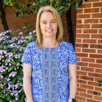 Headshot of Kristy Trent, a woman with blonde hair, smiling in a blue patterned shirt. She is standing outdoors in front of a brick wall and a flowering hydrangea bush.