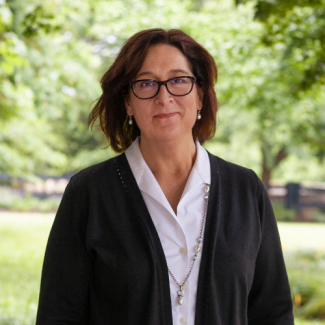 Laura Pennington, a woman with dark hair and glasses wearing a black cardigan over a white blouse and long silver necklace, smiling in an outdoor garden.