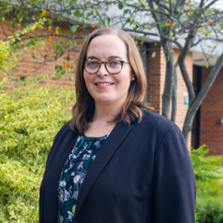 Portrait of Lindsey Honaker, a woman with brown hair and glasses, wearing a dark blazer over a floral top, standing outdoors.