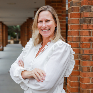 Portrait of Lizzie Goodpasture, a woman with blonde hair, smiling with arms crossed in front of a brick walkway.