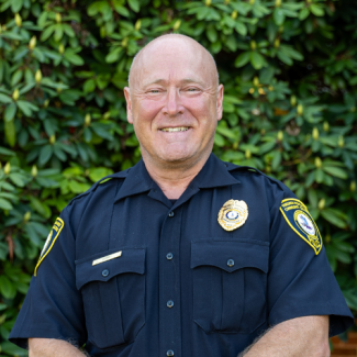 A man with a shaved head and a friendly smile wears a navy blue Virginia Highlands Community College police uniform. The uniform includes a gold badge on the chest, shoulder patches, and a gold nameplate. He stands against a backdrop of green rhododendron bushes.