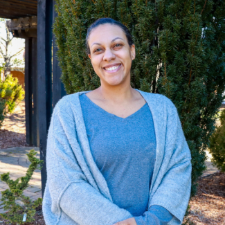 Professional headshot of Melanie Bush, a smiling woman with her hair pulled back, wearing a blue top and a light blue cardigan, standing in front of green shrubbery.