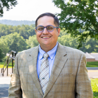 Headshot of Michael McBride smiling outdoors, wearing blue-rimmed glasses, a light blue dress shirt, and a plaid blazer.