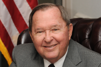 Close-up portrait of Bill Gatton, a smiling older man in a grey suit jacket and white shirt, seated in front of an American flag.