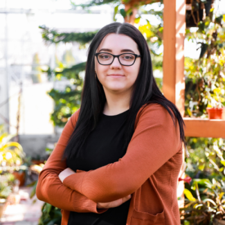 Professional headshot of Nicole Boothe, a woman with long dark hair and glasses, wearing a black top and an orange cardigan, standing in a sunlit greenhouse.