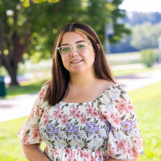 A smiling woman with brown hair, clear-framed glasses, and a floral smocked blouse outdoors in front of a blurred green background.