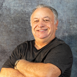 A man with grey hair and a warm smile poses with his arms crossed. He is wearing a dark grey, finely striped t-shirt and a silver watch. He stands against a grey, textured studio background.