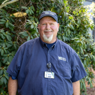 A man with a white goatee and a navy blue baseball cap smiles for a portrait. He is wearing a short-sleeved navy button-down shirt with a "Virginia Highlands Community College" logo on the pocket and a visible employee ID badge clipped to the front. He is standing in front of green foliage.