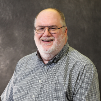 Robert May, a man with glasses and a white beard, wearing a grey plaid button-down shirt, smiling against a dark grey textured background.