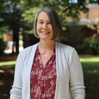 Robin Widener, a woman with a grey bob haircut wearing a maroon floral blouse and light grey cardigan, smiling in an outdoor setting.