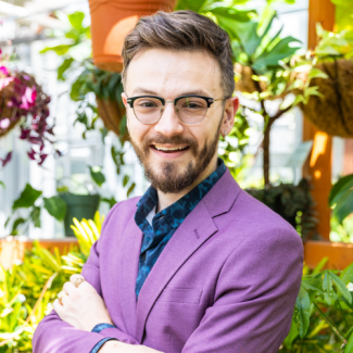 Headshot of Ryan Whittington smiling, wearing a purple jacket, glasses, and a patterned blue shirt, set against a background of lush greenhouse plants.