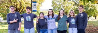 Seven students standing side-by-side with their arms around each other, smiling on a concrete campus walkway with autumn trees and a Virginia Highlands Community College banner in the background.