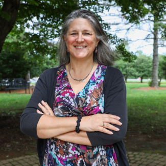 Professional headshot of Sandy Davis, a woman with long, graying hair, wearing a patterned dress and a black cardigan, standing outside with trees in the background.