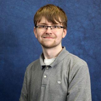 A young man with short brown hair, glasses, and a goatee smiles against a textured blue backdrop. He is wearing a grey polo shirt over a white undershirt.