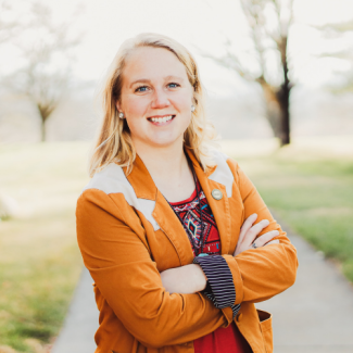 Shasta Wilson, a woman with blonde hair wearing an orange blazer over a red patterned shirt, standing with arms crossed in an outdoor park setting.