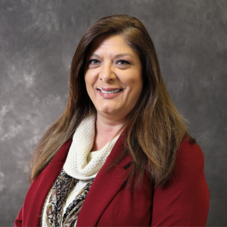Sherrie Phillips, a woman with long brown hair wearing a red blazer, white knit scarf, and leopard print undershirt, smiling against a grey studio backdrop.