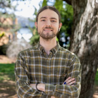 A young man with short brown hair and a smile poses with his arms crossed. He is wearing a black polo shirt featuring a small white logo on the chest. He stands outdoors in front of a green leafy bush.
