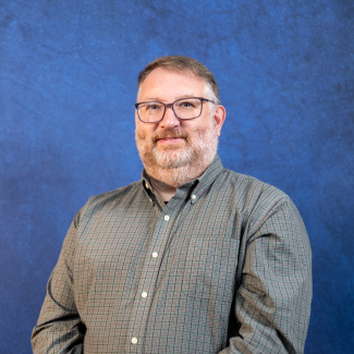 A professional headshot of a middle-aged man with short brown hair, a beard, and glasses, smiling and wearing a green checkered button-down shirt against a textured blue background.