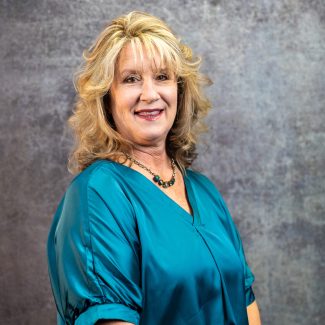 A woman with blonde, layered hair smiles. She is wearing a teal satin blouse and a dark beaded necklace. The background is a grey, textured studio wall.