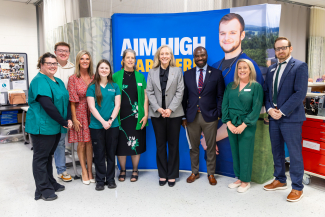 A group of nine people pose for a group photo inside a clinical simulation lab classroom at Virginia Highlands Community College. In the center stand Governor Abigail Spanberger in a grey blazer and black trousers, and Secretary of Health and Human Resources Marvin Figueroa in a navy blazer, red striped tie, and khaki trousers. They are flanked by college officials, faculty, staff, and nursing students in scrubs. They are standing in front of a blue backdrop banner that reads "AIM HIGH".