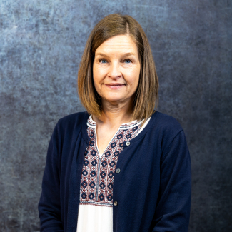A medium headshot of Beth Fairbanks looking directly at the camera with a kind expression. She is wearing a patterned white top and a navy cardigan. The lighting is bright and even, set against a dark, mottled grey-blue backdrop.