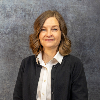 Headshot of Beth Fairbanks, a woman with shoulder-length brown hair, wearing a white collared shirt and a black cardigan, smiling against a dark grey textured background.