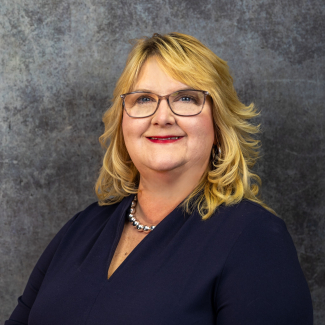 Headshot of Cindy Fields, a woman with blonde hair and glasses, wearing a navy blue top and a pearl necklace, smiling against a dark grey textured background.