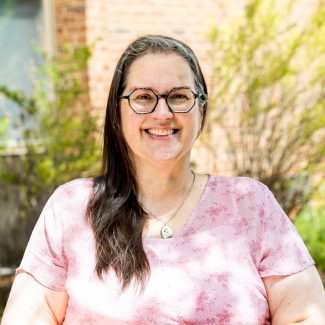 A medium close-up headshot of Dr. Patty Tymon smiling. She has long dark hair swept to one side, wears colorful geometric-framed glasses and a silver pendant necklace. She is wearing a pink floral patterned blouse against a soft-focus outdoor background of bricks and greenery.