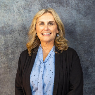 Professional headshot of Shannon Mutter smiling, wearing a blue polka-dot blouse and a black cardigan against a gray textured background.