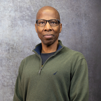 Headshot of Thomas Bryant, a man with a shaved head and black-rimmed glasses, wearing an olive green quarter-zip sweater against a textured gray background.