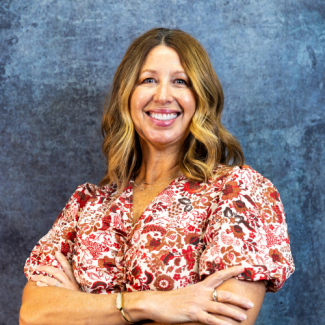 Professional headshot of Stacey Dorton smiling with her arms crossed, wearing a red and white floral blouse against a textured blue background.