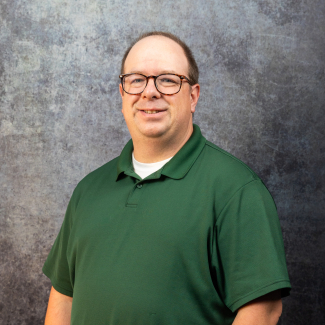 Josh Greenwell smiling at the camera, wearing tortoiseshell glasses, a white undershirt, and a dark green polo shirt in front of a mottled gray backdrop.