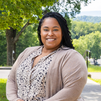 A medium shot of Alesha Russell, a smiling woman with long black braided hair, standing outdoors. She is wearing a beige cardigan over a cream-colored blouse with a black floral pattern. The background features green trees and a soft-focus view of distant hills.