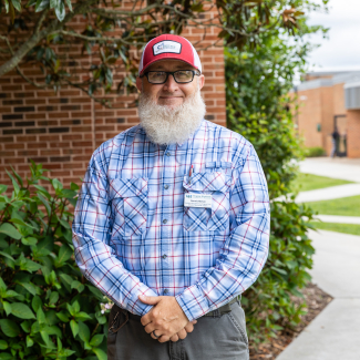 A smiling man with a full grey beard, glasses, and a red-and-white baseball cap standing outdoors with his hands clasped, wearing a blue and white plaid button-down shirt and grey pants.