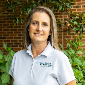 Headshot of Tamara Lasley smiling, wearing a white polo shirt with the Virginia Highlands Community College logo, standing in front of a brick wall and green foliage.