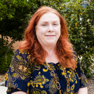 Teressa Wexler, a woman with long red hair wearing a blue and yellow paisley blouse and gold necklaces, smiling in front of green foliage.