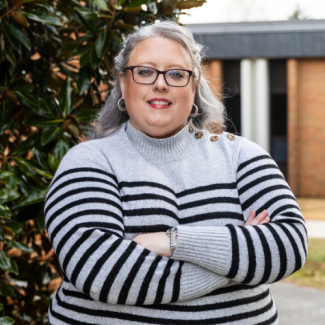 Professional headshot of Tiffanie Bishop, a woman with graying hair and glasses, wearing a gray and black striped sweater, standing outside with green foliage in the background.
