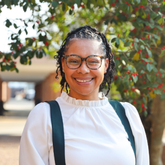 Professional headshot of Tiffany VanBuren, a woman with glasses and dark braided hair, smiling and wearing a white ruffled blouse with dark green overall-style straps against an outdoor background of trees with red berries.
