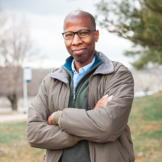 Tommy Bryant, wearing glasses and a jacket, standing outdoors with arms crossed.